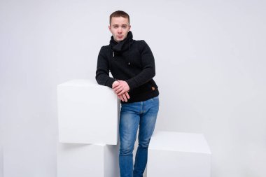 Kavramı studio bir küp üzerinde oturan ve konuşurken beyaz bir arka plan üzerinde genç bir adam portresi. Farklı pozlar ve kot pantolon ve kazak farklı duygular ile kamera karşısında oturuyor.