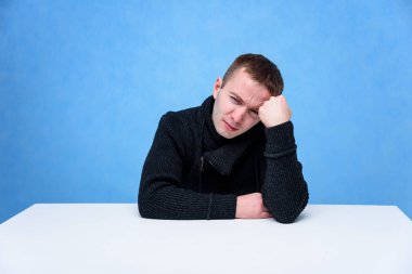 Mavi arka plan üzerinde bir küp üzerinde oturan genç bir adamın konsept stüdyo portresi. O farklı pozlar kamera karşısında oturur ve kot pantolon ve bir kazak farklı duygular ile.