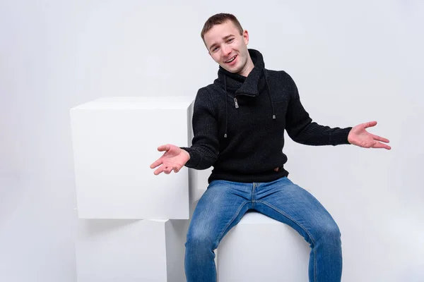 概念工作室肖像的一个年轻人在白色的背景坐在一个立方体和说话 他穿着不同的姿势坐在镜头对面 穿着牛仔裤和毛衣 有不同的情绪 — 图库照片