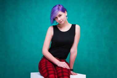 Buraya bakın - Yeşil bir arka plan üzerinde mor saçlı güzel bir kızın konsept portresi bir küp üzerinde oturan ve mal reklamını. O stüdyoda çeşitli pozlar kameranın hemen önünde.