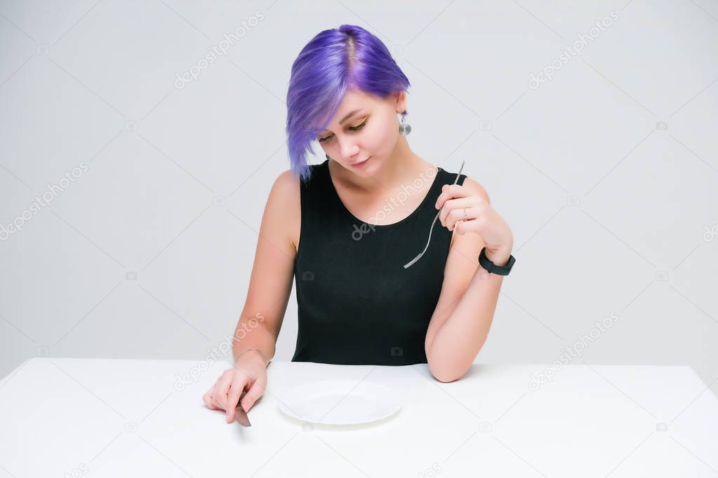 Fork, plate, knife - Concept portrait of a beautiful girl with purple hair on a white background sitting at a table with dining tools. She is in front of the camera in various poses in the studio.