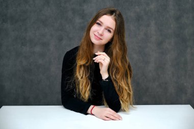 Kavramı bir hoş samimi ve iyimser mutlu esmer kız güzel uzun saçlı masada oturan gri bir arka plan üzerinde portresi. O tişört siyah bir gülümseyen ve konuşuyor.