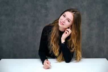 Kavramı bir hoş samimi ve iyimser mutlu esmer kız güzel uzun saçlı masada oturan gri bir arka plan üzerinde portresi. O tişört siyah bir gülümseyen ve konuşuyor.