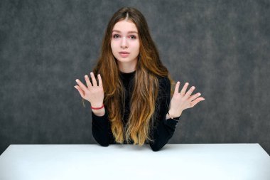 Kavramı bir hoş samimi ve iyimser mutlu esmer kız güzel uzun saçlı masada oturan gri bir arka plan üzerinde portresi. O tişört siyah bir gülümseyen ve konuşuyor.