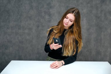 Kavram. Mutlu brunet kız güzel uzun saçlı bir telefonla bir masada oturan hoş bir dostu Müdürü portresi. O bir siyah T-shirt gri bir arka planda gülümseyen içinde.