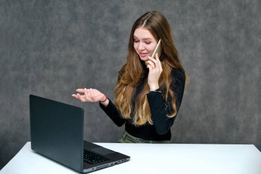 Mutlu brunet kız güzel uzun saçlı bir telefon ve dizüstü bilgisayar ile bir masada oturan hoş bir dostu Müdürü kavramı portresi. O bir siyah T-shirt gri bir arka planda gülümseyen içinde.