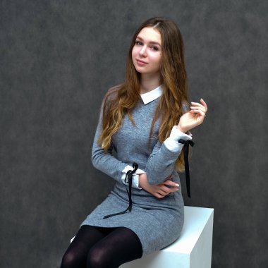 Bir küp üzerinde güzel uzun saçlı mutlu bir esmer kız hoş bir dostu Müdürü kavramı portresi. Tam büyüme gri bir arka plan karşı gülümseyen bir gri elbiseli duruyor.