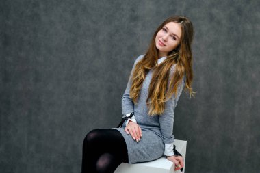 Bir küp üzerinde güzel uzun saçlı mutlu bir esmer kız hoş bir dostu Müdürü kavramı portresi. Tam büyüme gri bir arka plan karşı gülümseyen bir gri elbiseli duruyor.