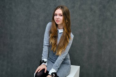 Bir küp üzerinde güzel uzun saçlı mutlu bir esmer kız hoş bir dostu Müdürü kavramı portresi. Tam büyüme gri bir arka plan karşı gülümseyen bir gri elbiseli duruyor.