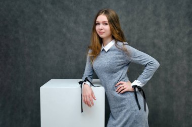 Bir küp üzerinde güzel uzun saçlı mutlu bir esmer kız hoş bir dostu Müdürü kavramı portresi. Tam büyüme gri bir arka plan karşı gülümseyen bir gri elbiseli duruyor.