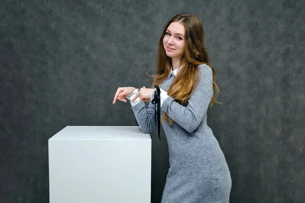 Concept portrait of a pleasant friendly manager of a happy brunette girl with beautiful long hair on a cube. She is standing in a gray dress smiling against a gray background in full growth.