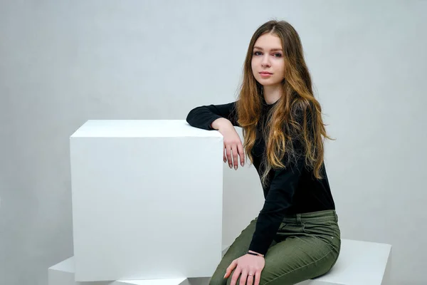 Concept portrait of a brunette girl, sitting on a white background in a black t-shirt on a white cube in studio. She sits right in front of the camera in various poses with different emotions.