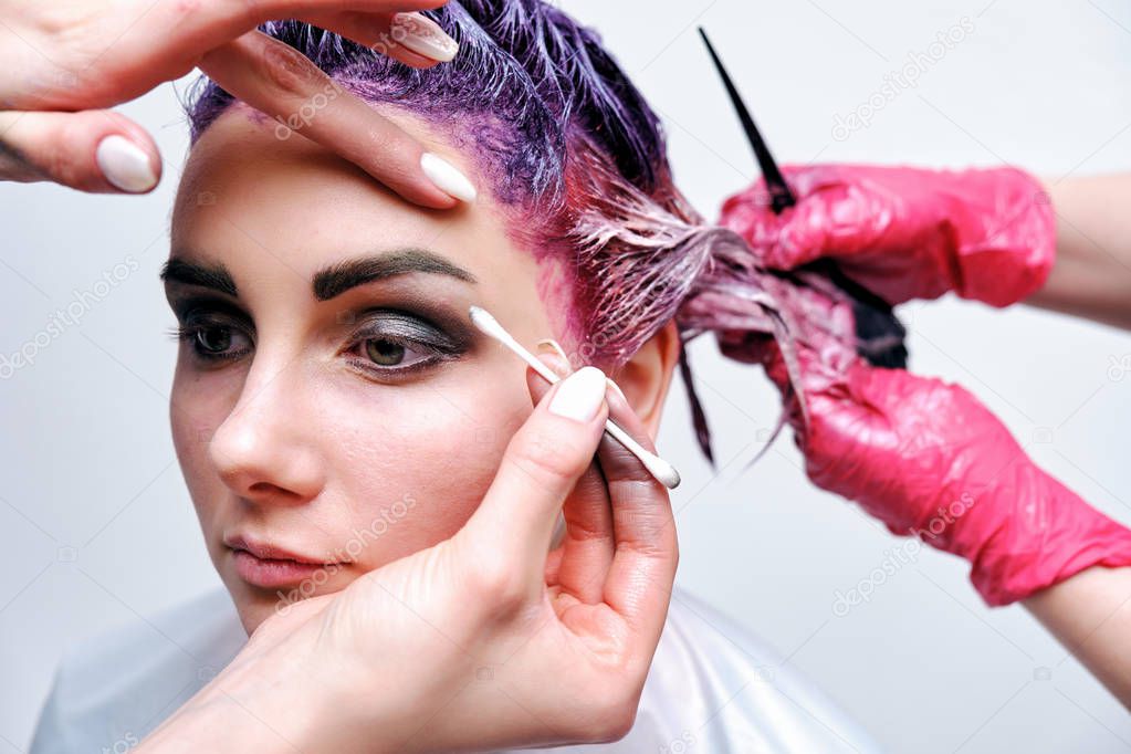 Beautiful girl with violet hair on white background master in make-up. Visible hand of the master and his makeup. The girl is in the middle of the frame and sits on a chair.