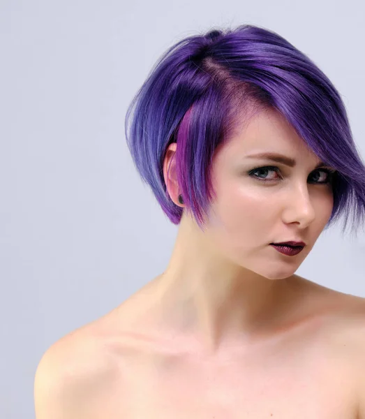 A beautiful, sexy girl with purple hair and a short haircut sits in the middle of the photo with a white background and grimaces. She\'s naked and visible shoulders.