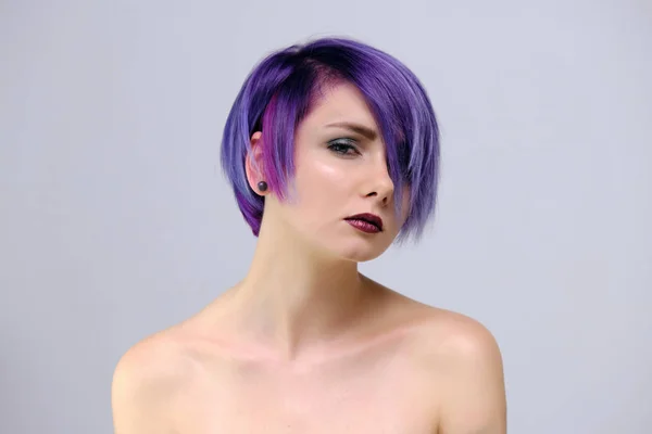 A beautiful, sexy girl with purple hair and a short haircut sits in the middle of the photo with a white background and grimaces. She\'s naked and visible shoulders.