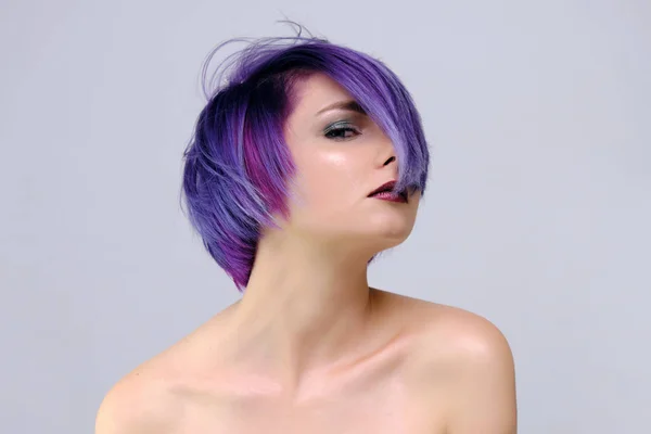 A beautiful, sexy girl with purple hair and a short haircut sits in the middle of the photo with a white background and grimaces. She\'s naked and visible shoulders.