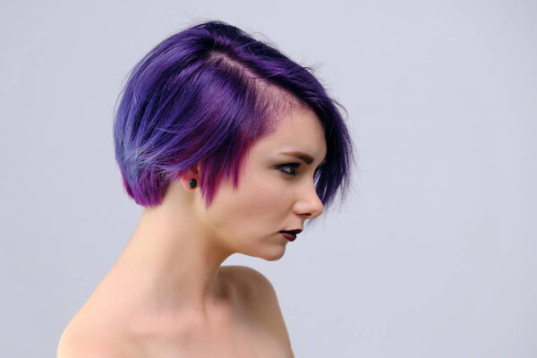 A beautiful, sexy girl with purple hair and a short haircut sits in the middle of the photo with a white background and grimaces. She's naked and visible shoulders.
