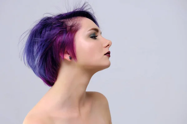 A beautiful, sexy girl with purple hair and a short haircut sits in the middle of the photo with a white background and grimaces. She\'s naked and visible shoulders.