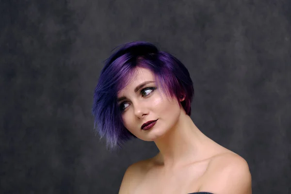 A beautiful, sexy girl with purple hair and a short haircut sits in the middle of the photo with a gray background and grimaces. She\'s naked and visible shoulders.