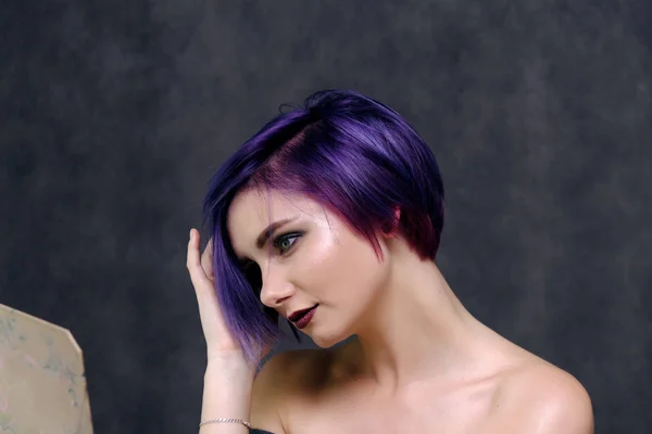 A beautiful, sexy girl with purple hair and a short haircut sits in the middle of the photo with a gray background and grimaces. She\'s naked and visible shoulders.