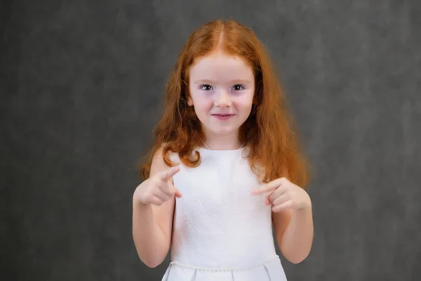 Concept portrait of a cute pretty child girl with red hair on a gray background smiling and talking — Stock Photo, Image