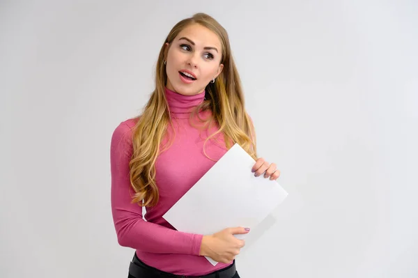 Ritratto ravvicinato di una bella ragazza bionda con lunghi capelli ricci in piedi in studio su uno sfondo bianco con emozioni in pose diverse in un maglione rosa con una cartella in mano . — Foto Stock