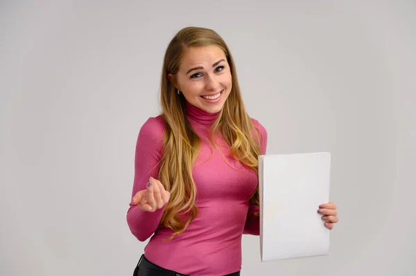 Retrato de cerca de una hermosa chica rubia con el pelo largo y rizado de pie en el estudio sobre un fondo blanco con emociones en diferentes poses en un suéter rosa con una carpeta en las manos . —  Fotos de Stock