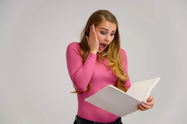 Gros plan portrait d'une jolie fille blonde aux longs cheveux bouclés debout en studio sur fond blanc avec des émotions dans différentes poses dans un pull rose avec un dossier dans ses mains . — Photo