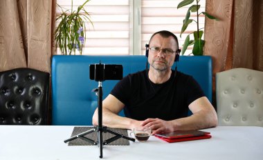 Siyah tişörtlü bir model kameranın tam karşısında bekliyor. Evde akıllı telefon ve tabletle video konferansı düzenlerken kulaklık ve gözlüklü yetişkin bir adamın portresi bekliyor..