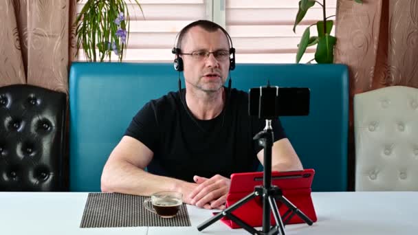 Blanke jongeman met een baard in een zwart T-shirt zittend op een bank en met een draadloze headset — Stockvideo