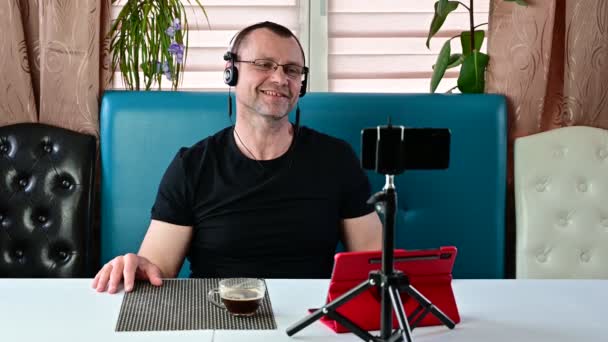 Blanke man met een baard in een zwart T-shirt zittend op een bank en met een draadloze headset — Stockvideo