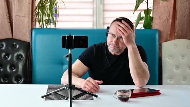 Caucasian young man in a black T-shirt sits on a sofa and puts on a headset — Stock Video