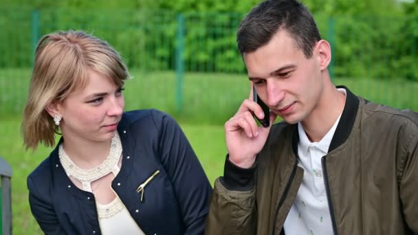 Un hombre está hablando por teléfono. Pareja joven discutiendo mientras se sienta en un banco en un parque. El concepto de conflicto familiar . — Vídeos de Stock