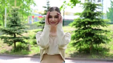 Modaya uygun bir kadın. Mükemmel makyajlı, gülümseyen beyaz bir kız güneşin altındaki bir şehir parkında duruyor..