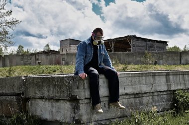 Bir binanın harabelerinin fotoğrafı. Koruyucu maskeli bir adam güneşli bir havada harabelerde oturur.
