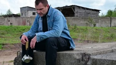Terk edilmiş binanın arka planında bir adam. Bir adam elinde maskeyle harabelerde oturuyor.