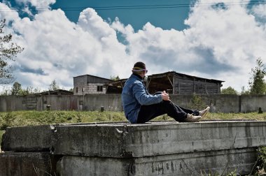 Bir binanın harabelerinin fotoğrafı. Koruyucu maskeli bir adam güneşli bir havada harabelerde oturur.