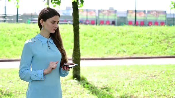 Hübsche Frau in blauer Bluse mit Handy in der Hand bei sonnigem Wetter. — Stockvideo