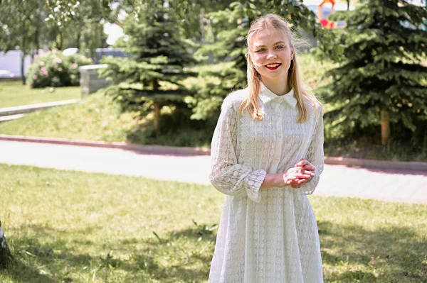 Ağaçların arka planındaki beyaz model güneşli havada duruyor. Parkta gülümseyen güzel sarışın bir kızın portresi.