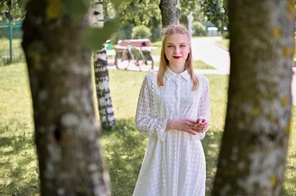 Ağaçların arka planındaki beyaz model güneşli havada duruyor. Parkta gülümseyen güzel sarışın bir kızın portresi.