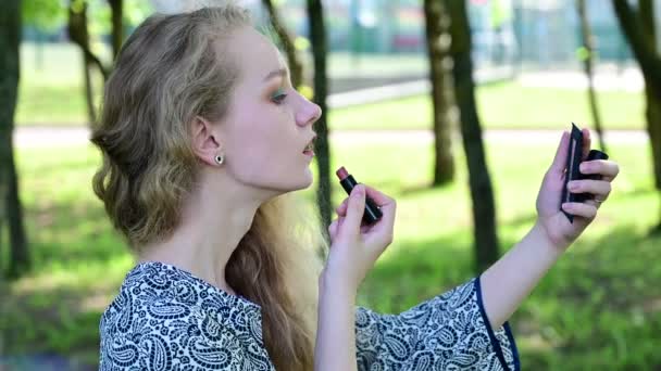 Vidéo caucasienne blonde fille peint les lèvres avec du rouge à lèvres dans le parc. — Video