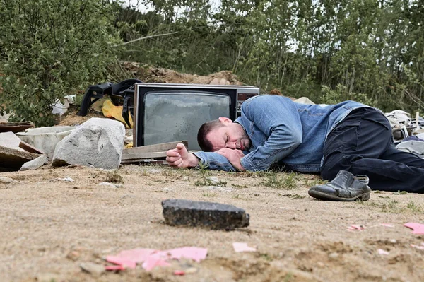 Photo of a homeless man on the background of trash and garbage outdoors. Model is lying on the ground and looking at the camera