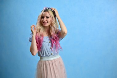 Kafasında çelenk olan beyaz bir kadının yatay portresi. Stüdyoda mavi arka planda gülümseyen sarı saçlı model.