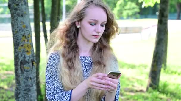 Aantrekkelijke jonge blanke vrouw met lang krullend haar in een park maakt gebruik van een telefoon. — Stockvideo