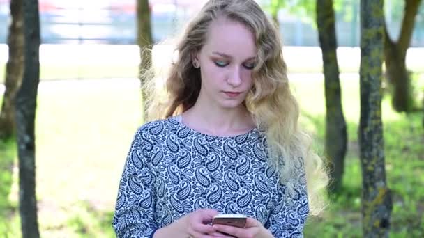 Een mooi meisje van Europees uiterlijk met lang krullend haar gebruikt een telefoon in een park. — Stockvideo