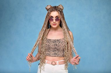 Mavi arka planda kameranın önünde duran güzel bir model. Stüdyoda uzun saçlı afro saçlı, gözlüklü beyaz bir kadının portresi.