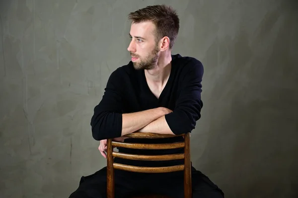 Stylish vertical studio portrait of a caucasian young man with a beard sitting on a chair on a gray background — Stock Photo, Image