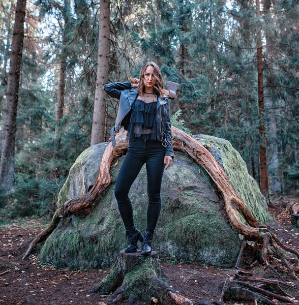 Молодая красивая женщина в кожаной куртке с топором в лесу — стоковое фото