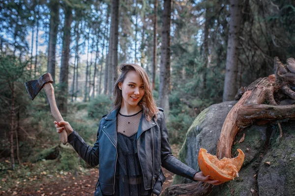 Young beautiful woman in leather jacket with pumpkin and axe in a forest — Stock Photo, Image