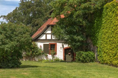 Yarı ahşap mimarisi olan küçük bir peri masalı evi ve çalılıkların ve gül çalılarının arasında bir ağacın altında kırmızı kiremitli bir çatı.